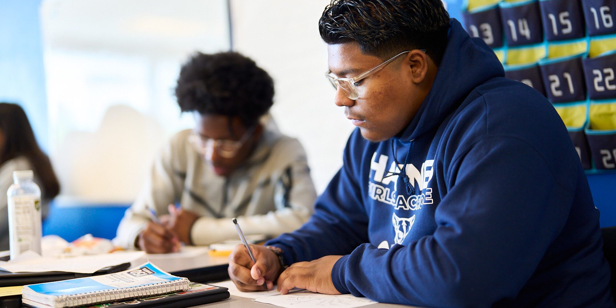 A photo of a male Latino high school student writing on paper, with a Black male student also working in the background.